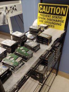 hard drives stacked in a data recovery laboratory. A sign in the background reads: Caution, prevent static spark discharge. Use grounding devices.