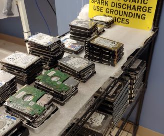 hard drives stacked in a data recovery laboratory. A sign in the background reads: Caution, prevent static spark discharge. Use grounding devices.
